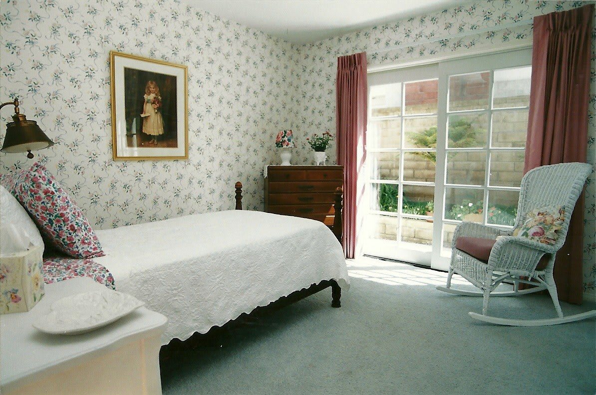A cozy bedroom with floral wallpaper, a large bed, and sunlight streaming through glass doors.