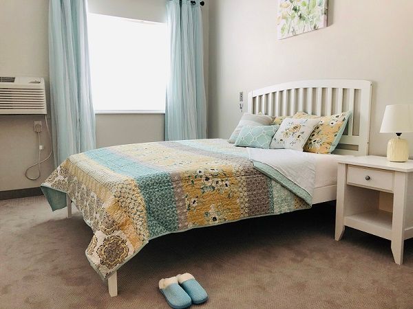 A cozy bedroom featuring a neatly made bed with a colorful quilt, complemented by matching pillows and light blue curtains. Slippers are placed beside the bed.