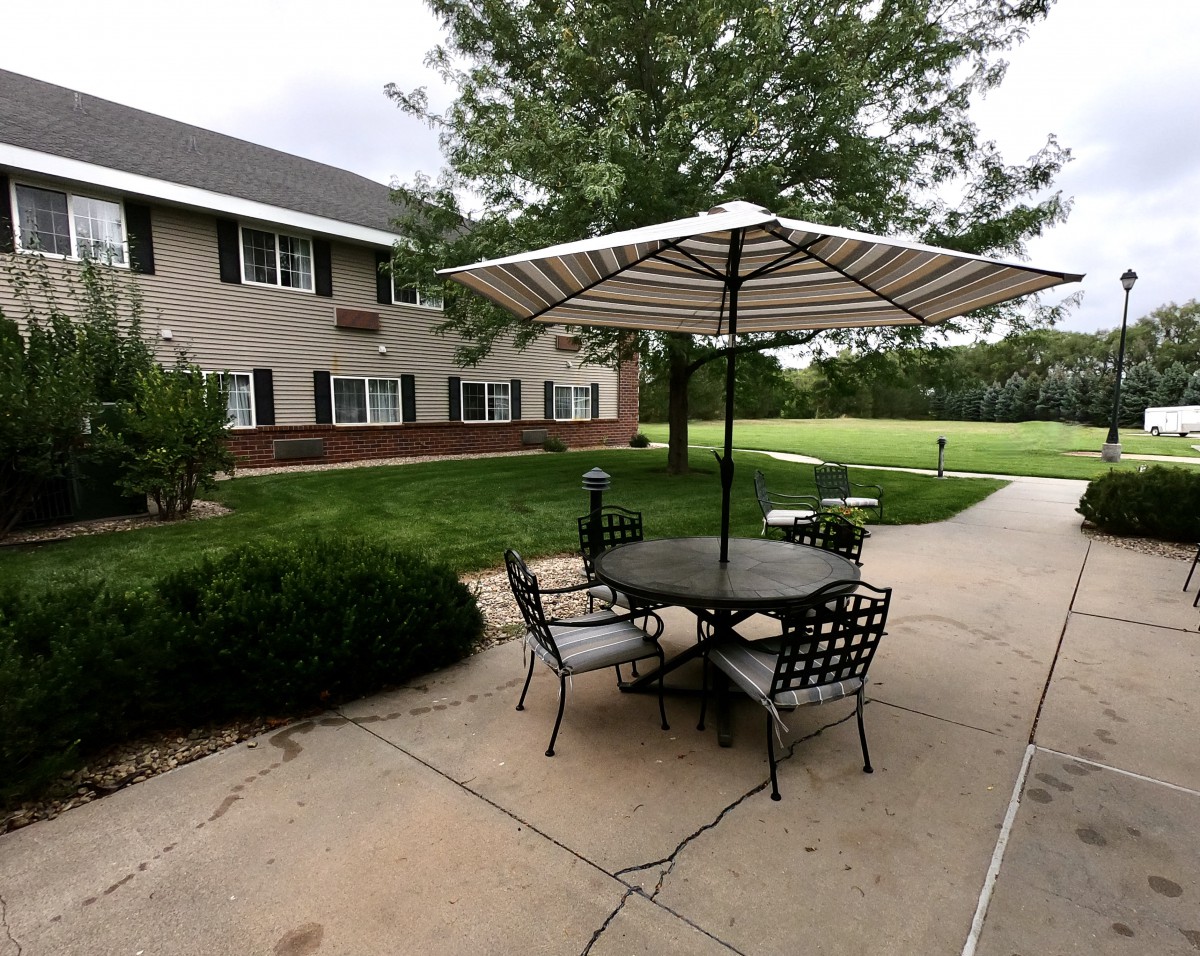A serene outdoor patio with a table and chairs under an umbrella, adjacent to a building surrounded by greenery.