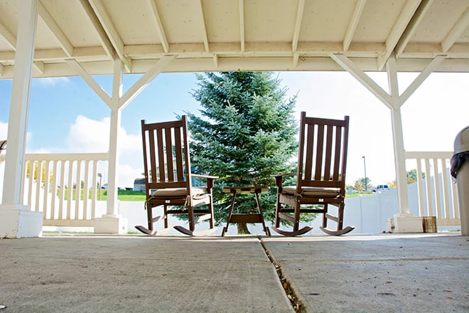 A pair of wooden rocking chairs on a covered porch facing a lush green tree.
