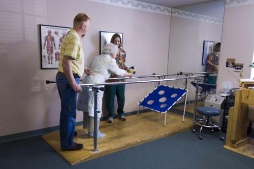 An elderly person receives assistance during a physical therapy session using parallel bars.
