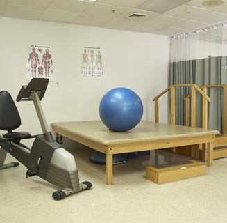 A rehabilitation room equipped with a stationary bike, an exercise ball, and a therapy platform.