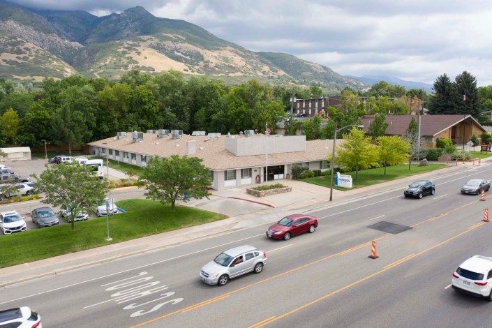 A view of a road with cars passing by, set against a backdrop of mountains and surrounded by trees on a cloudy day.