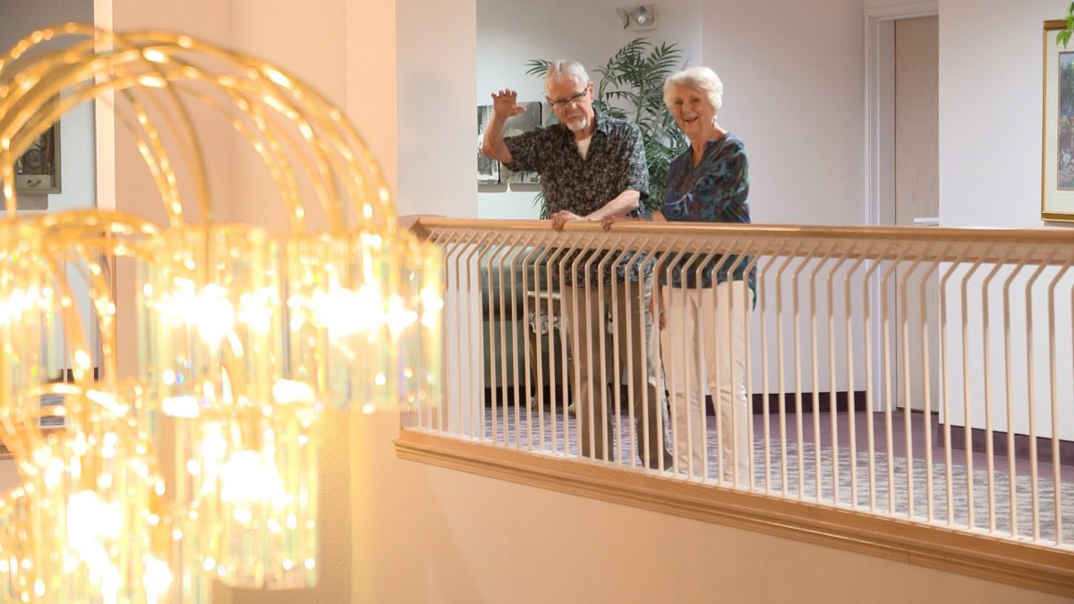 An elderly couple waves from a balcony inside a well-lit building.