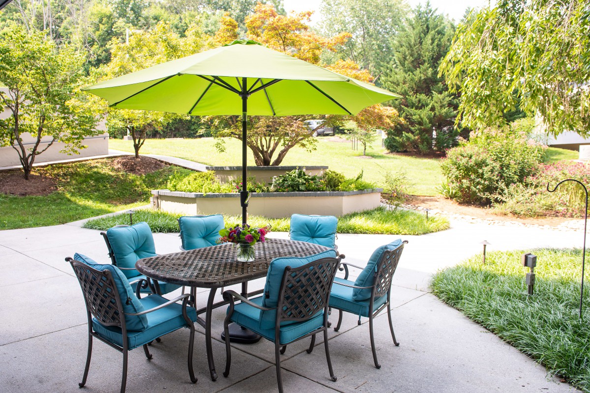 A cozy outdoor patio area with a table, chairs, and a bright green umbrella set against a lush garden backdrop.