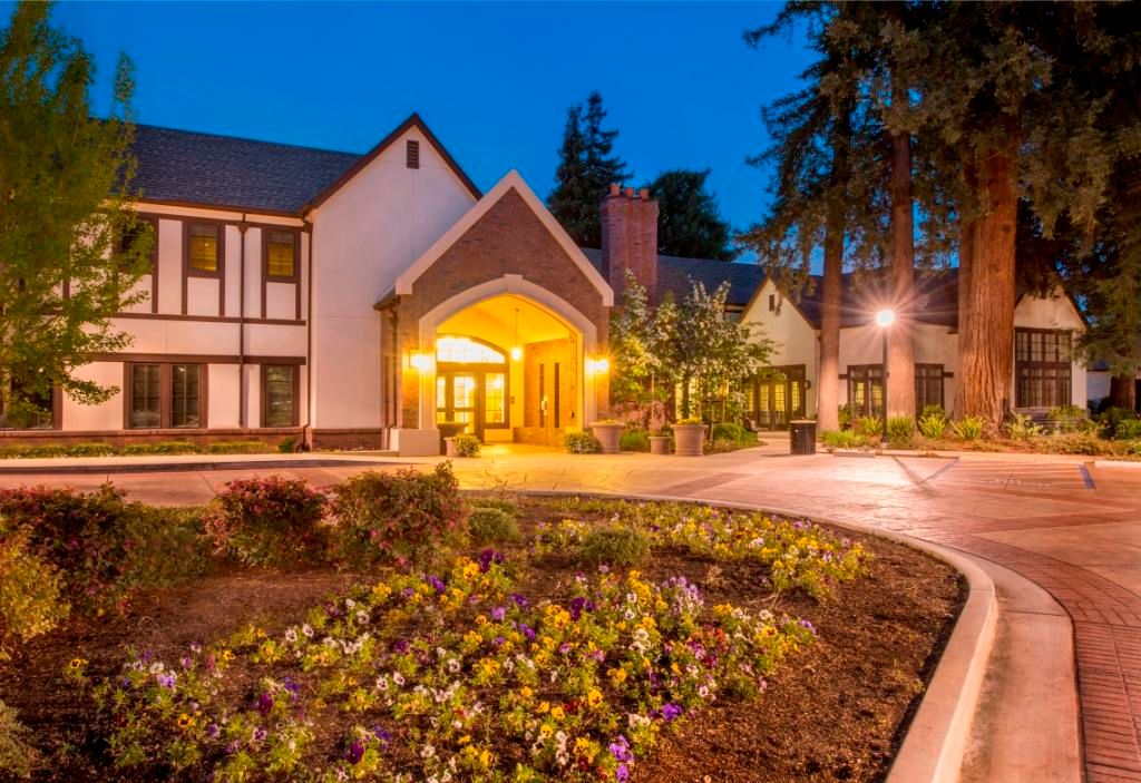 A charming house with a beautifully lit entrance, surrounded by gardens and trees during the evening.