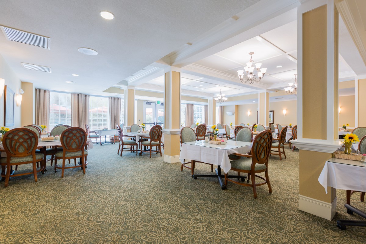 A spacious and elegantly decorated dining room with tables and chairs arranged neatly, featuring chandeliers and large windows providing natural light.