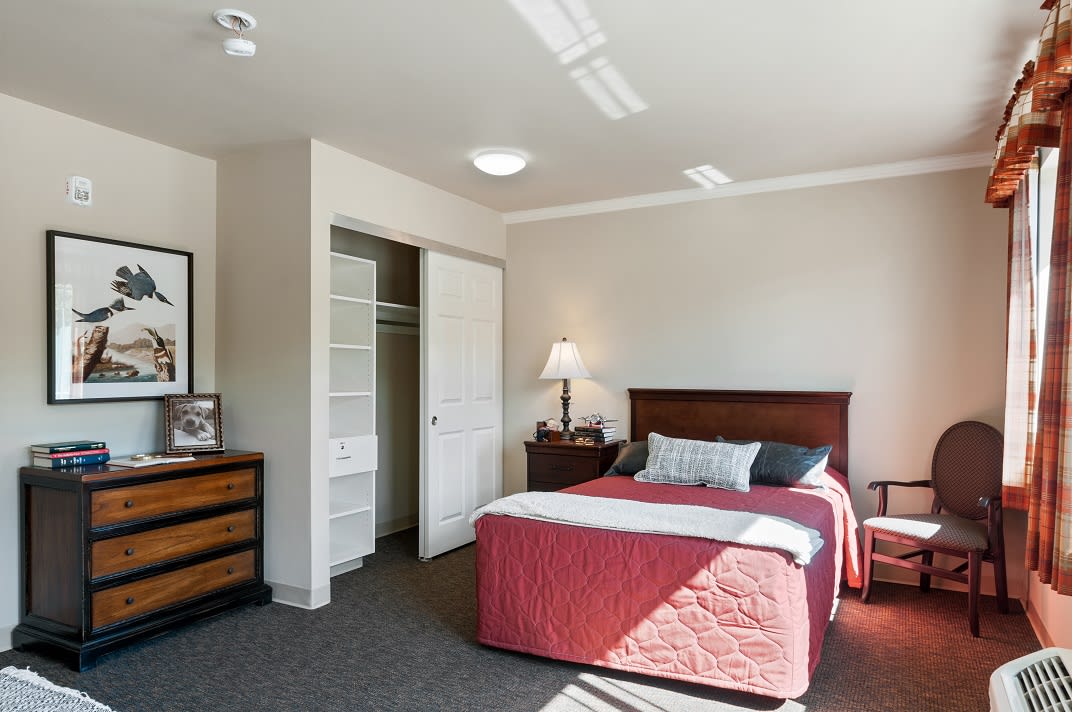 A cozy bedroom with a neatly made bed, a lamp on the nightstand, and an open closet.