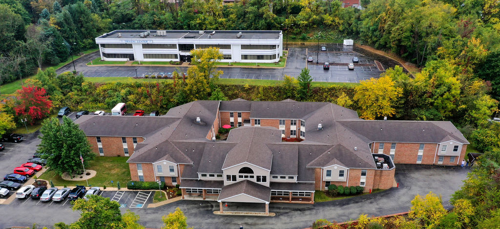 Aerial view of a healthcare facility surrounded by autumn foliage, with a parking lot and another building in the background.