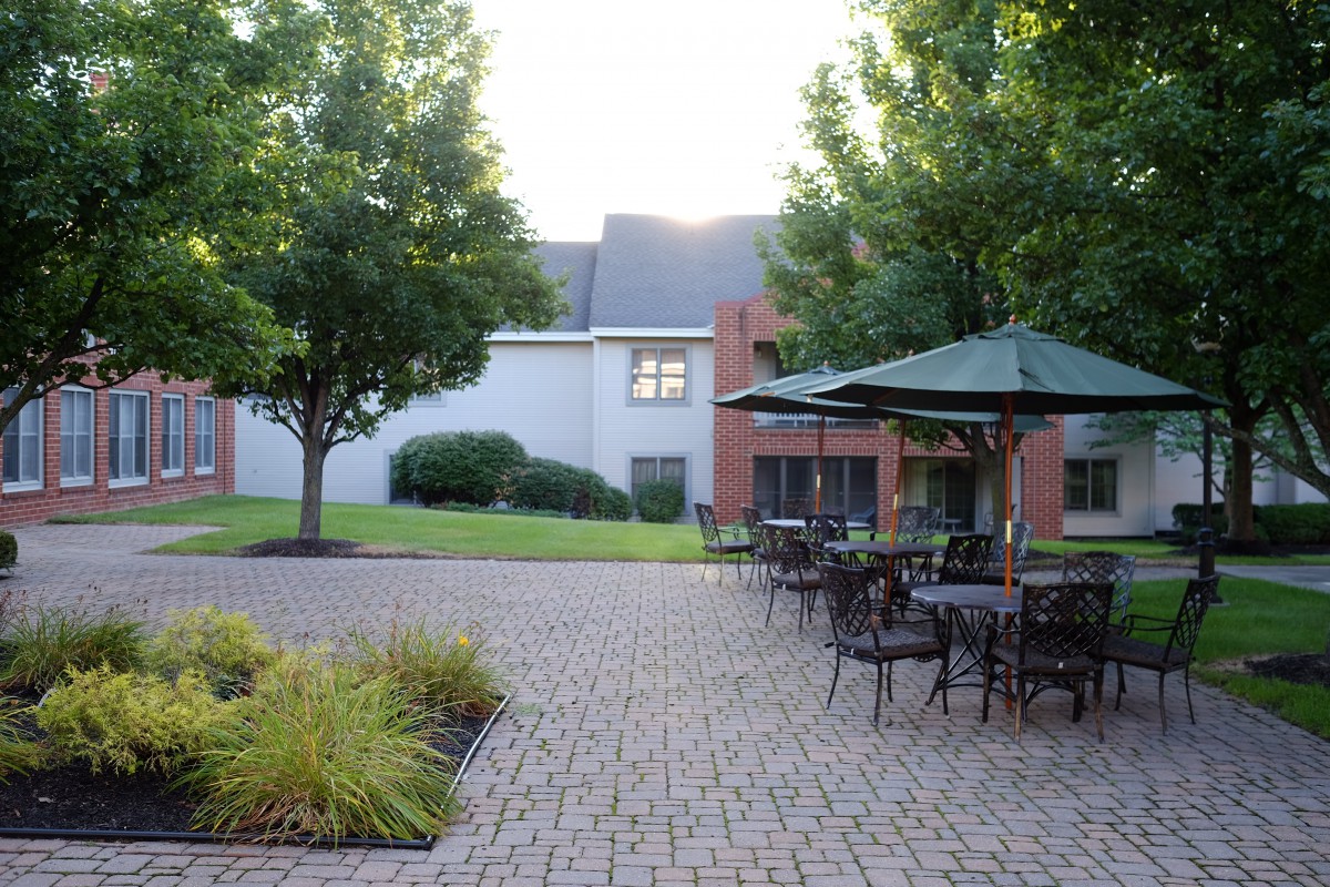 A peaceful courtyard with tables and umbrellas surrounded by trees and a brick building.