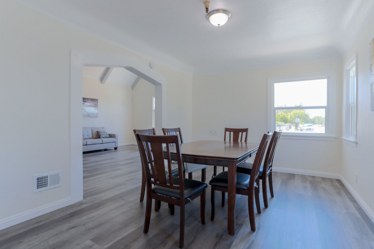 A minimalist dining room with a wooden table and chairs, featuring bright natural lighting.