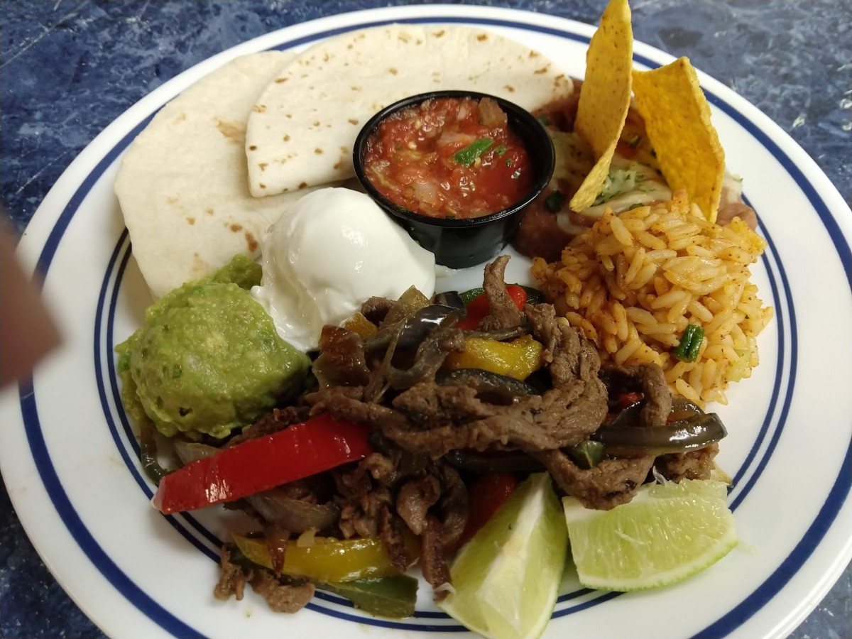 A colorful plate of Mexican cuisine featuring beef fajitas, accompanied by tortillas, rice, guacamole, sour cream, salsa, and lime wedges.