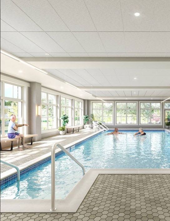 A bright indoor swimming pool area with large windows allowing for natural light and a couple of people sitting by the edge.