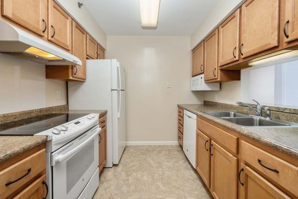 A narrow kitchen featuring wooden cabinets, white appliances, and a tile floor.