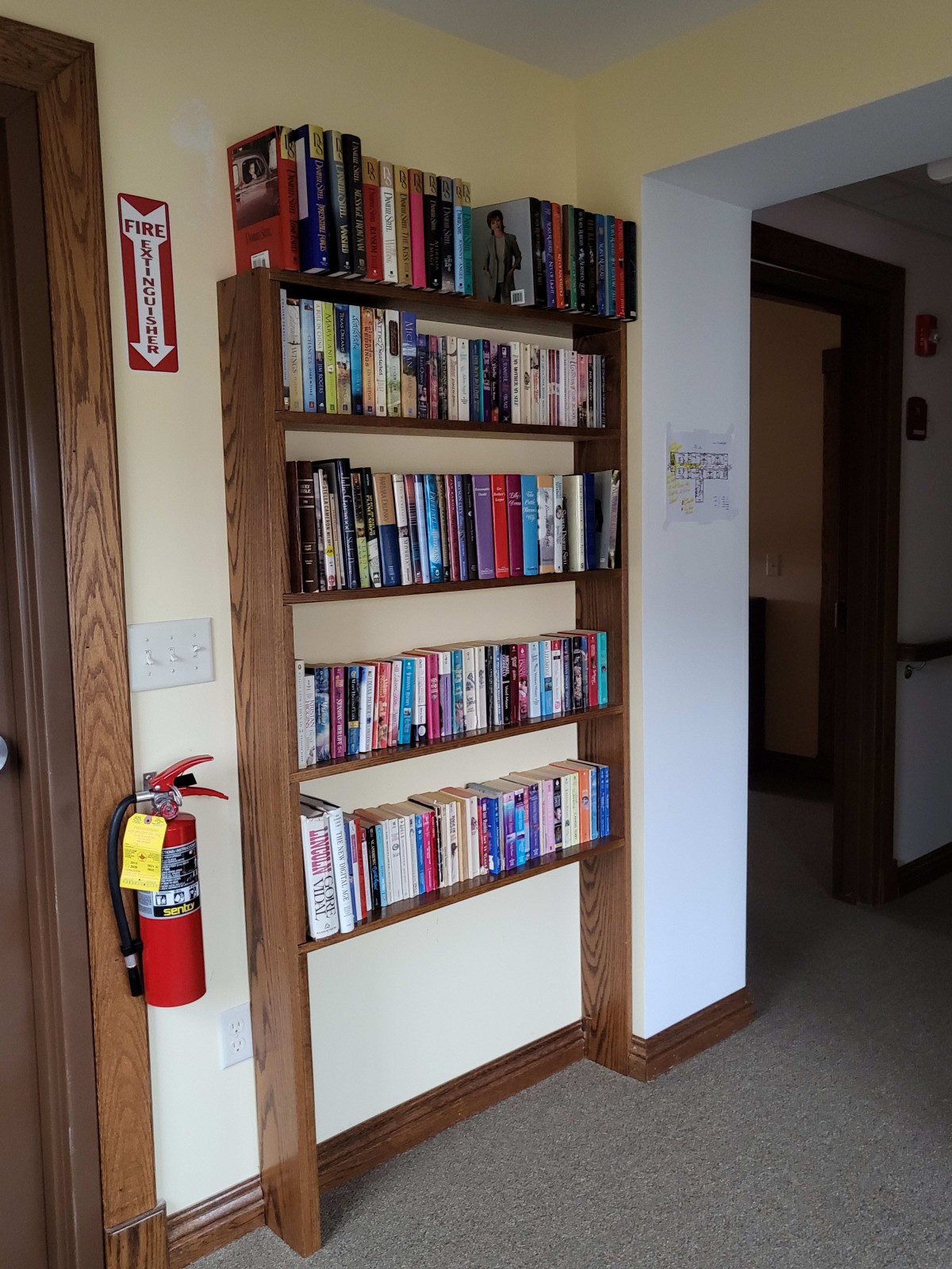 A hallway with a wall-mounted bookshelf filled with books alongside a fire extinguisher.
