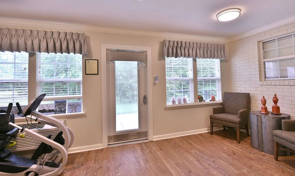 A cozy living room with wooden flooring, featuring a door, large windows with curtains, and comfortable seating.