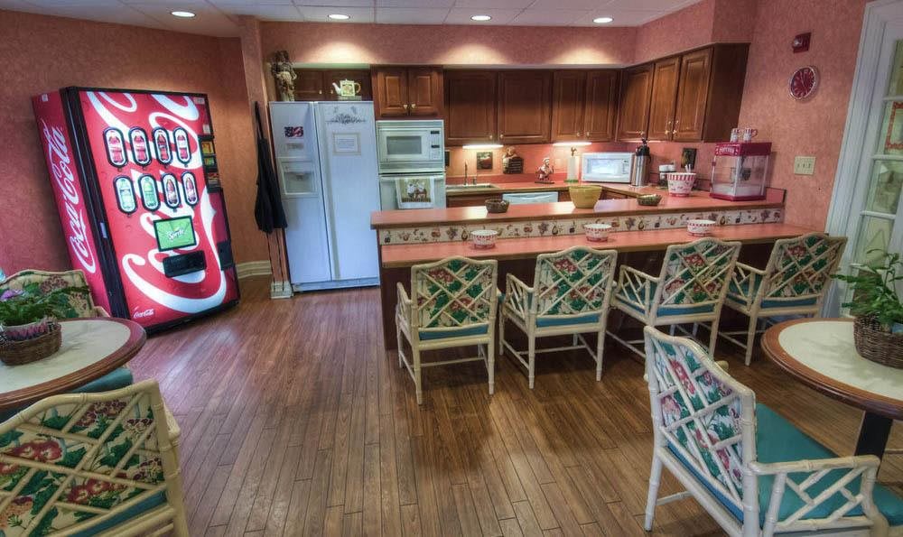 A spacious kitchen with a bar counter and chairs, featuring wooden cabinets and flooring, along with a refrigerator and a vending machine.