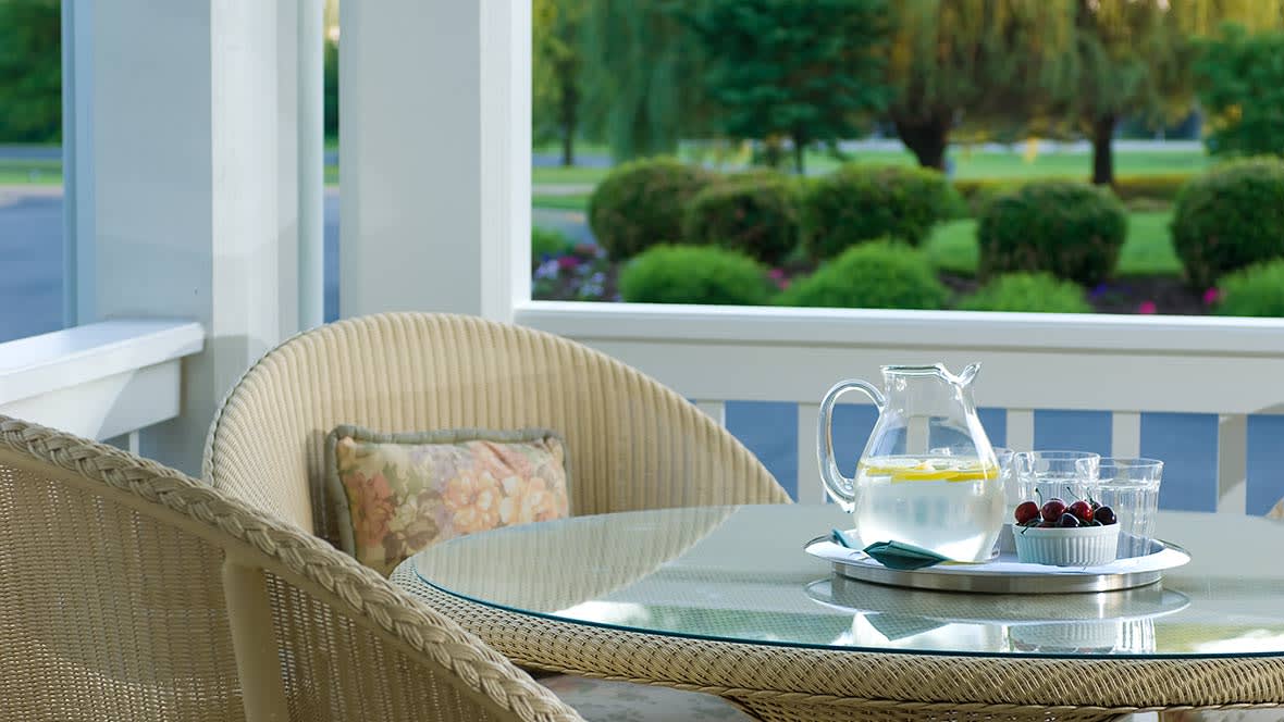 A cozy patio setting with a glass table, wicker chair, and a pitcher of lemon water.