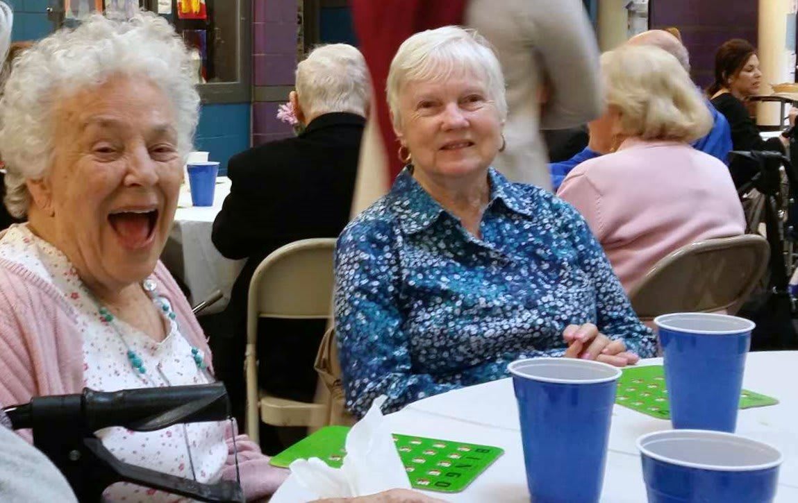 Elderly individuals enjoying a social gathering indoors, seated around tables with cups and conversation.