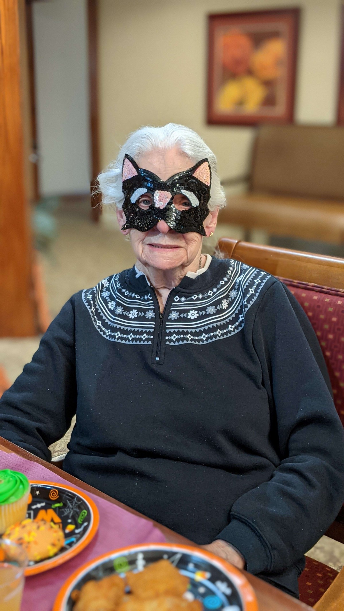 An elderly person wearing a cat mask, seated indoors.