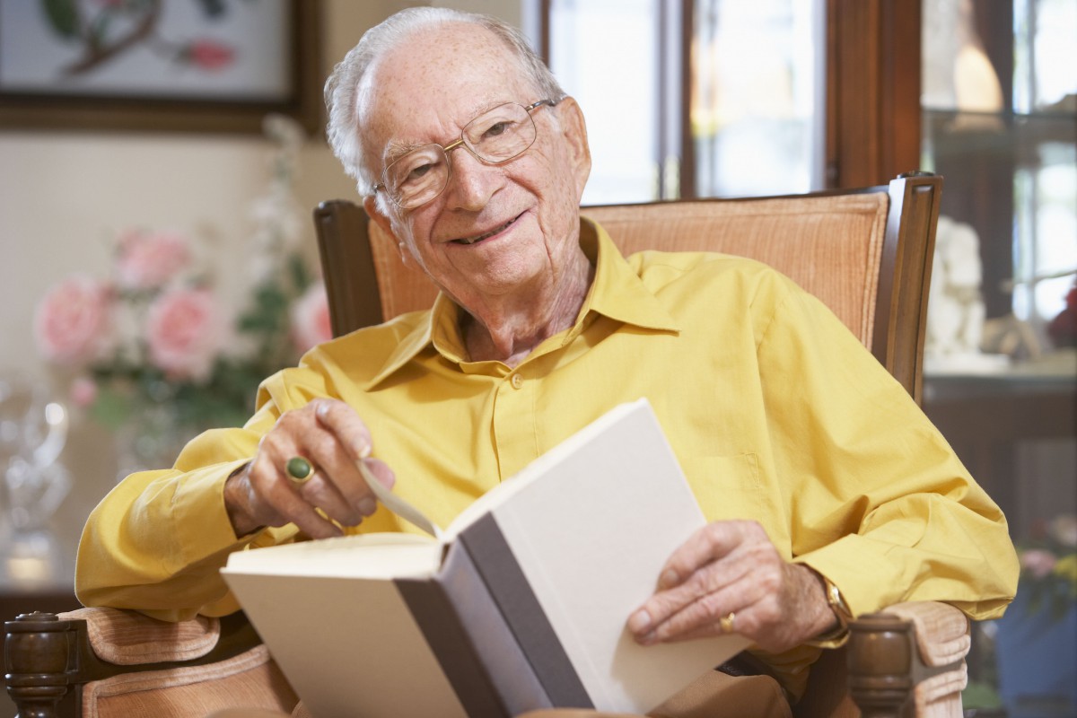 An elderly man sitting in a chair and smiling while holding an open book.