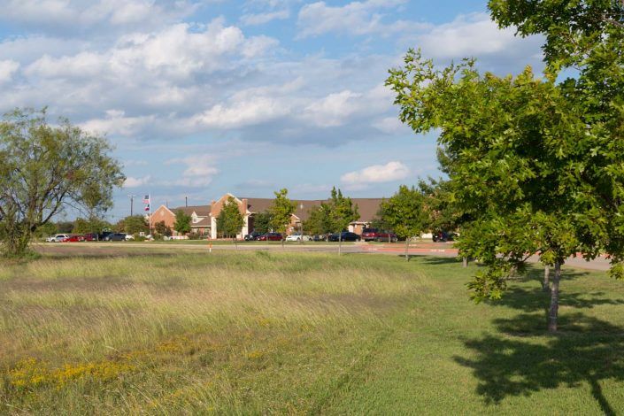 A scenic view of a grassy field with trees and buildings under a partly cloudy sky.