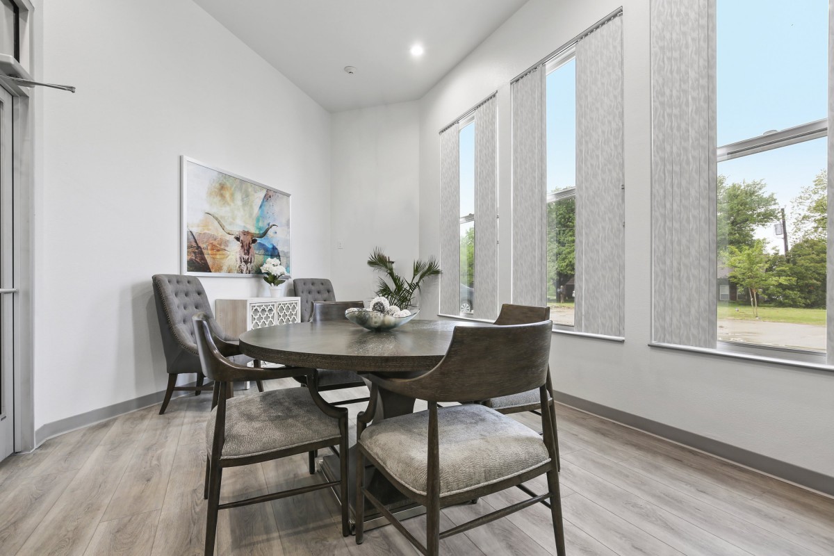 A modern dining room with a round wooden table, surrounded by upholstered chairs and large windows allowing natural light. Minimalist decor includes artwork and an indoor plant.
