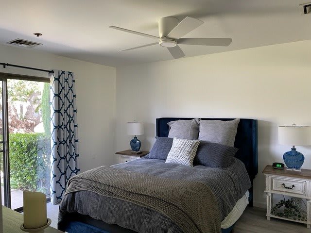 A cozy bedroom with a neatly made bed, featuring gray bedding and multiple pillows. A nightstand with a lamp sits beside the bed, and patterned curtains adorn the window.
