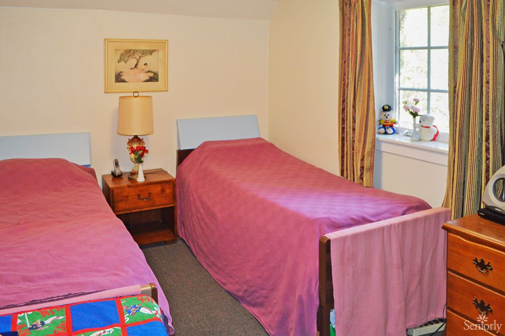 A cozy bedroom featuring twin beds with pink covers, a nightstand with a lamp, and a window with curtains.