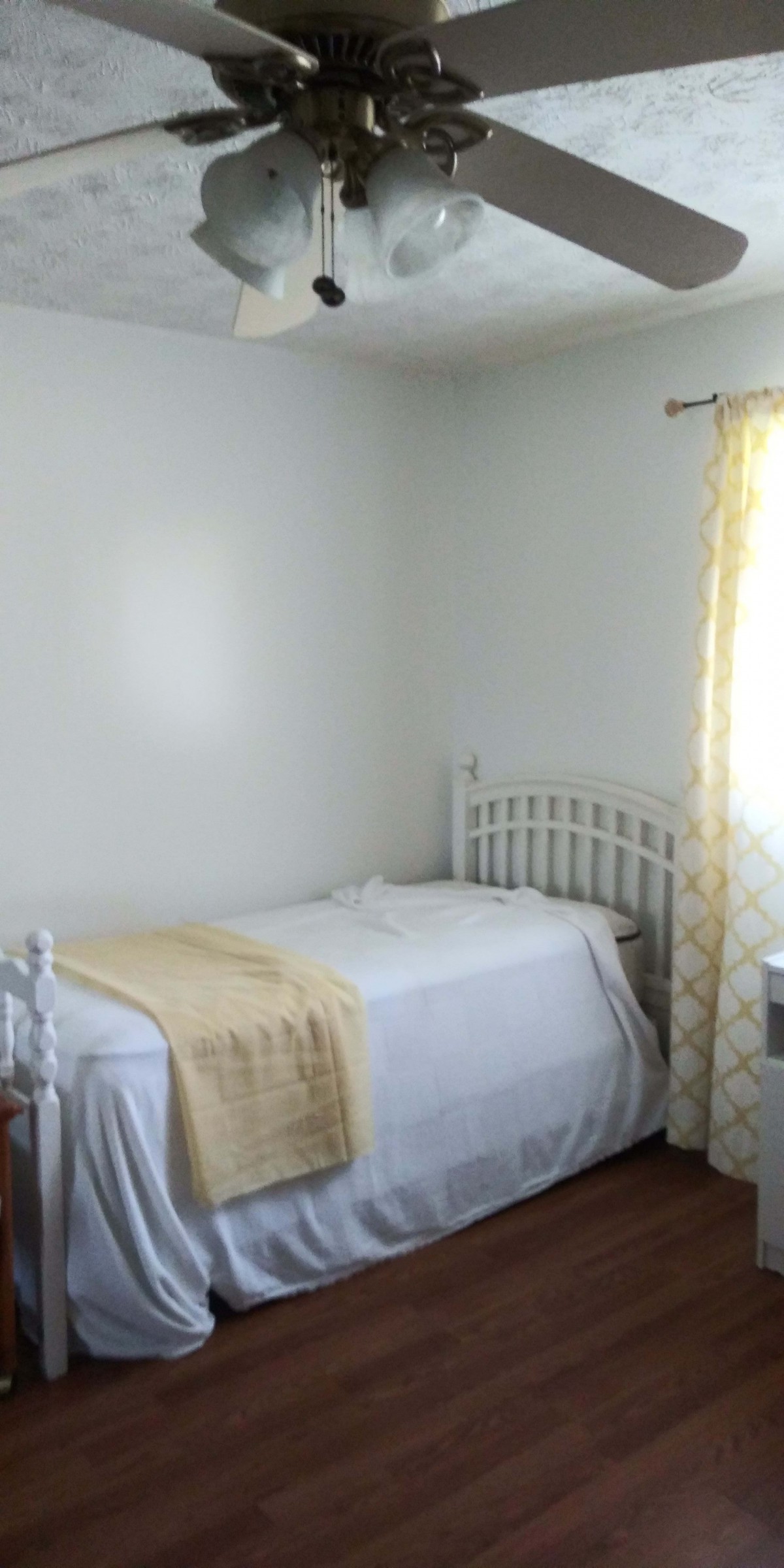 A minimalistic bedroom with a single bed and white bedding, softly illuminated by natural light from a window.
