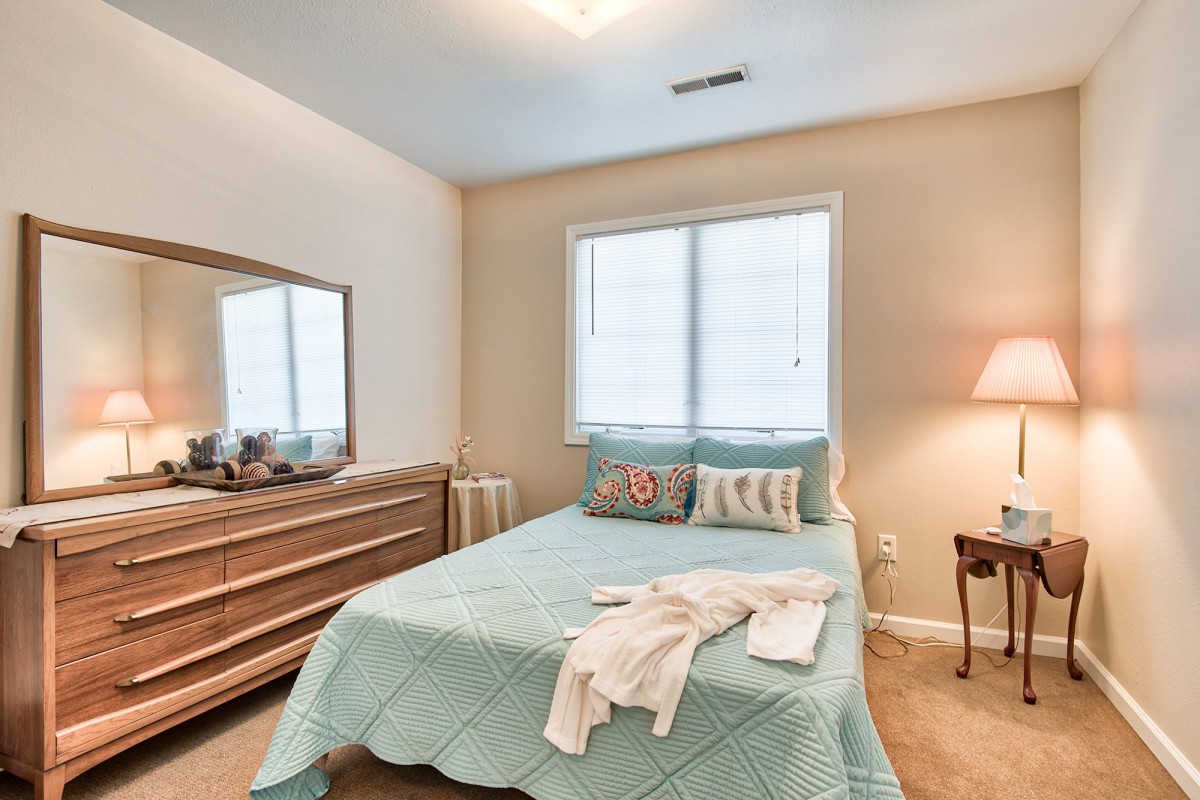 A cozy bedroom featuring a neatly made bed with a light blue comforter, a wooden dresser, and a soft lamp beside the window.