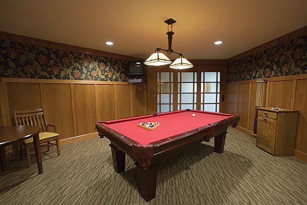 A billiard room featuring a red-felt pool table with overhead lighting and wood-paneled walls.