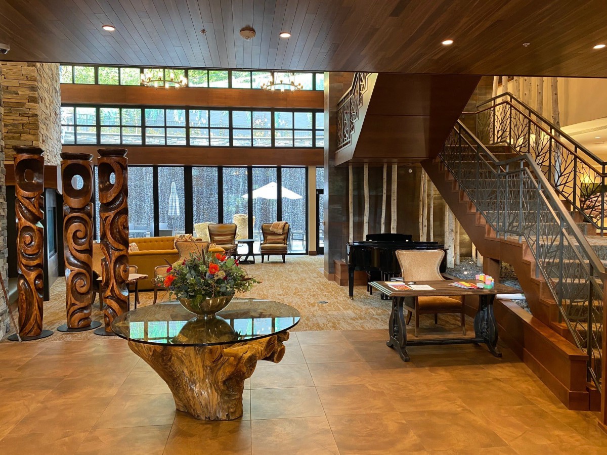 A spacious and modern lobby featuring rustic wooden furniture, a staircase, and large glass windows allowing natural light to fill the space.
