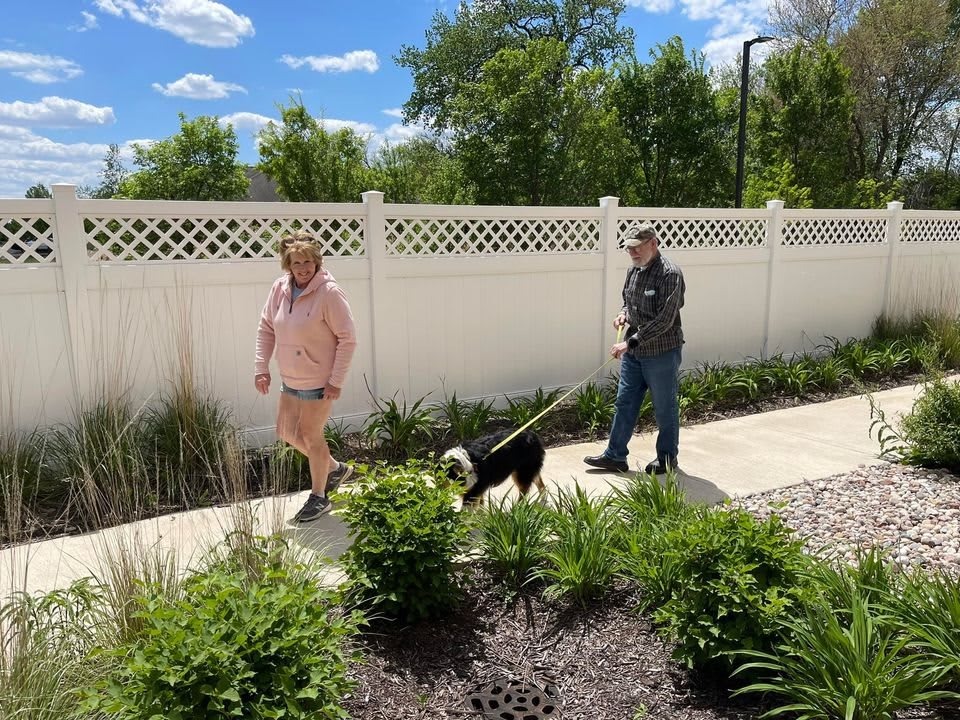 A couple walking their dog along a sunny sidewalk next to a white fence.