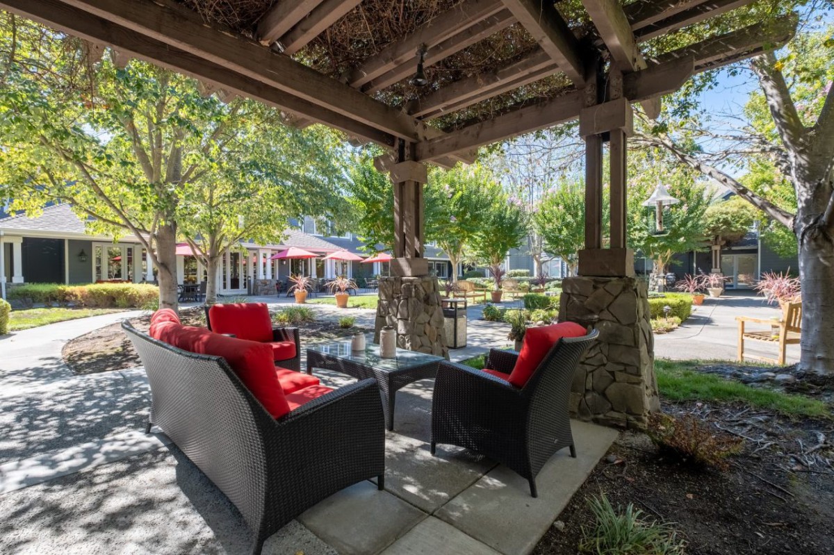 A cozy outdoor patio with wicker furniture and red cushions, under a wooden gazebo overlooking a garden.