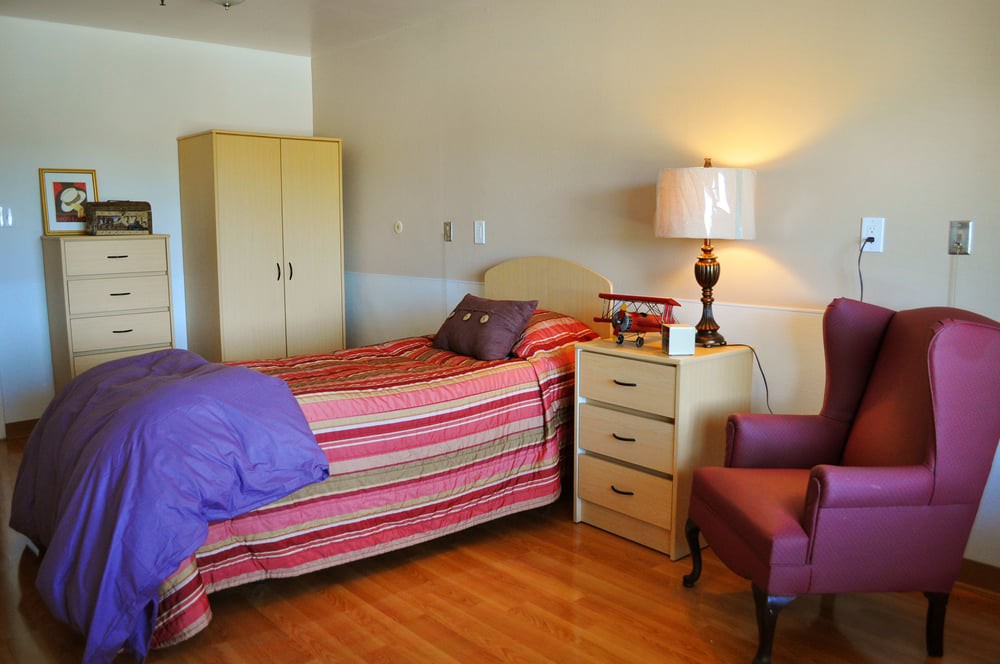 A cozy bedroom featuring a single bed with striped bedding, a wooden nightstand with a lamp, an armchair, and a wardrobe.