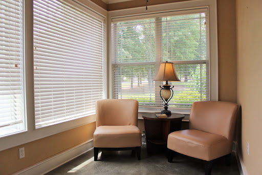 A cozy corner of a living room featuring two chairs, a table lamp, and natural light filtering through the blinds.
