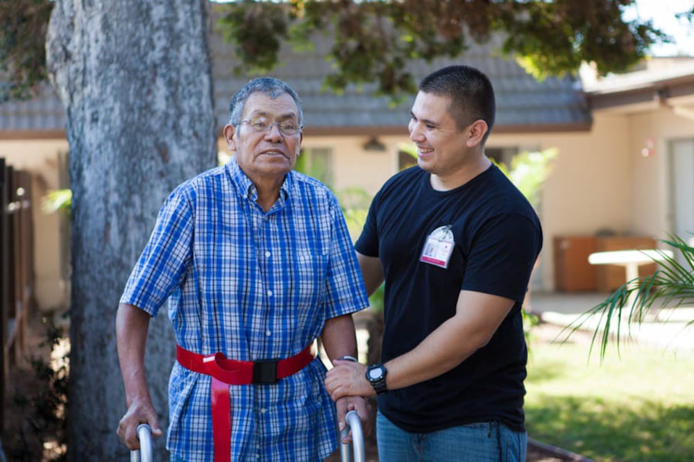 A caregiver assists an elderly man with a walker outside.
