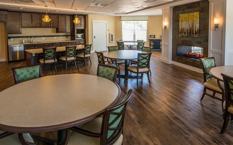 A spacious dining area with round tables and chairs, featuring a kitchen in the background and wooden flooring.