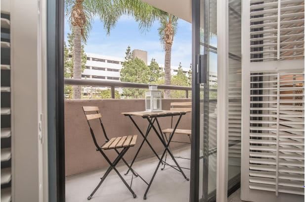 A cozy balcony with a small table and two chairs, overlooking palm trees and nearby buildings.
