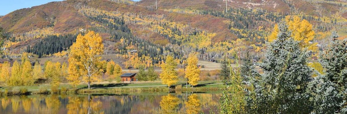 A serene autumn landscape with a cabin by a lake, surrounded by colorful trees and reflected in the water.