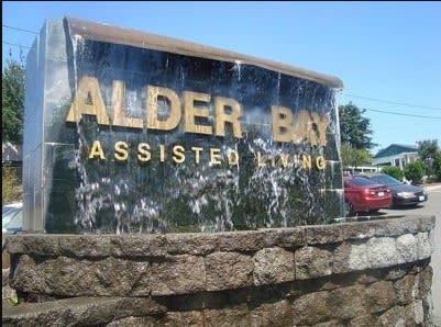 A sign for Alder Bay Assisted Living located outdoors, with a water feature in the background.