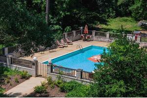 A serene outdoor swimming pool surrounded by lush greenery and a fence, with lounge chairs and an umbrella nearby.