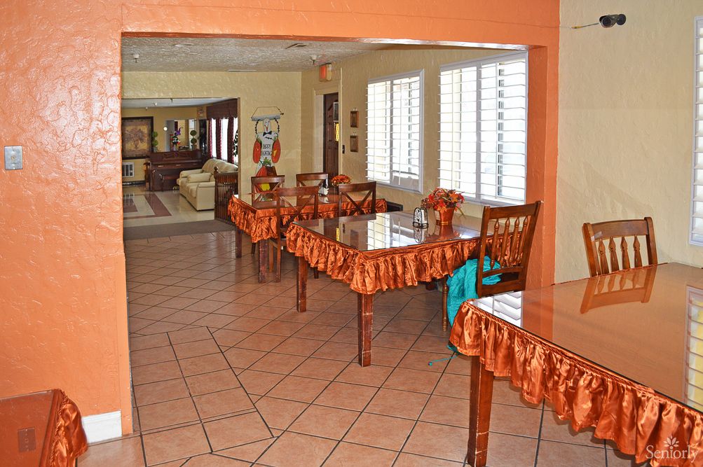 A cozy dining area with several wooden tables and chairs, decorated with orange tablecloths, set on a tiled floor with natural light streaming in through the windows.