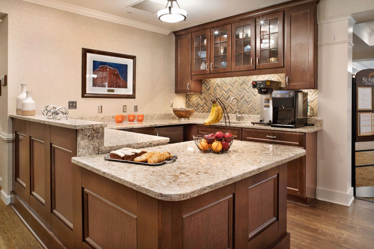 A modern kitchen with wooden cabinets and marble countertops, featuring a central island with a fruit bowl and coffee maker.