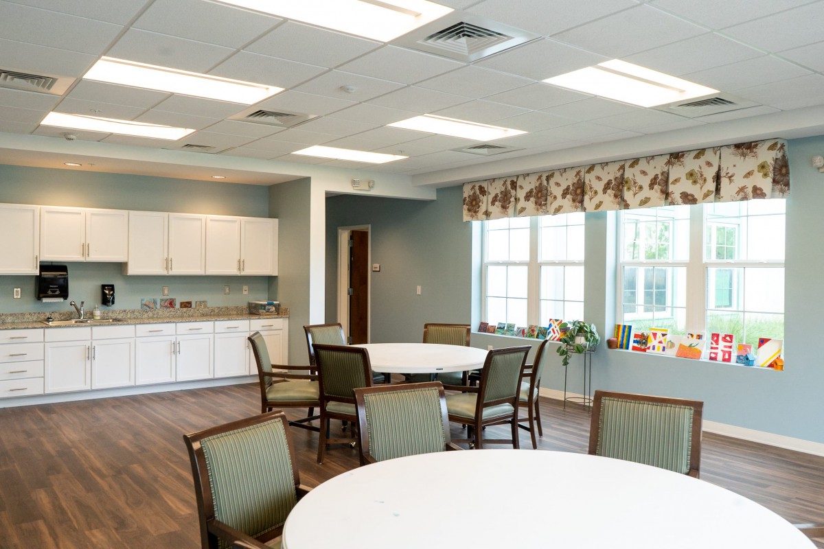 A bright and spacious dining area with round tables and chairs, featuring a kitchenette in the background.