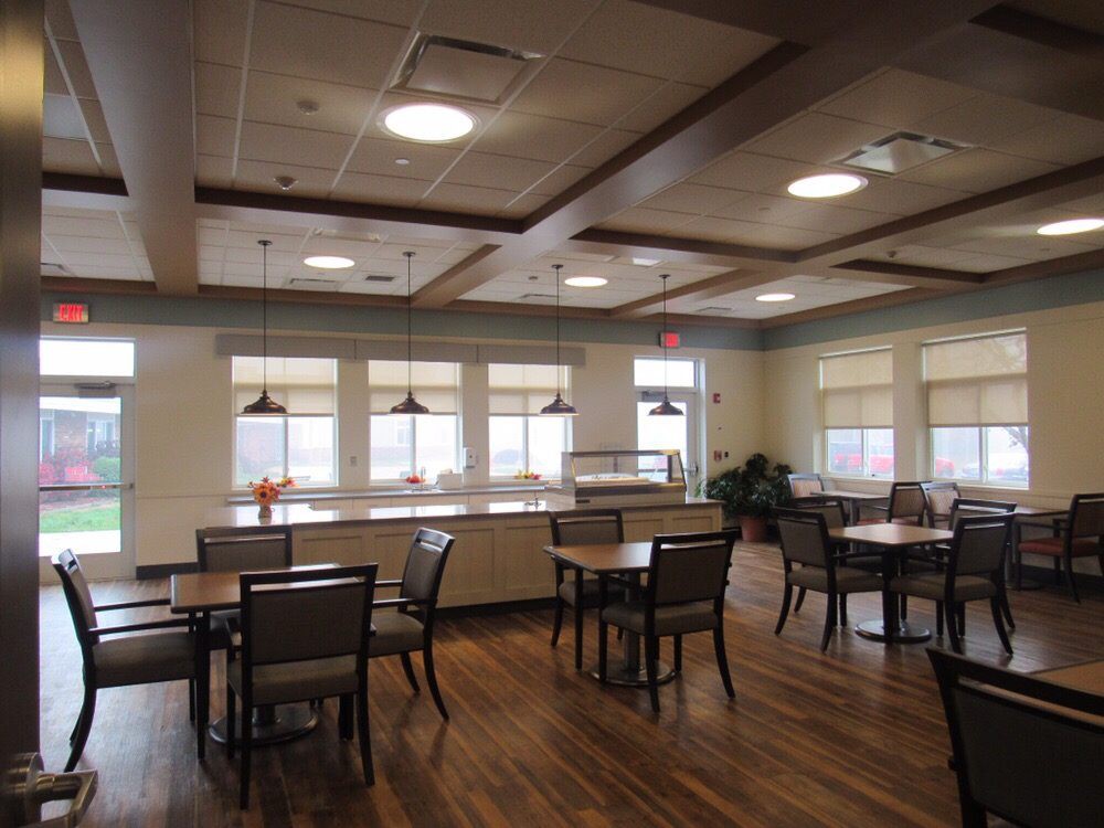 A spacious dining area with wooden floors, tables, and chairs, featuring large windows and ceiling lights.