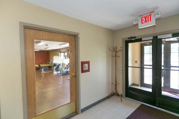A hallway with a glass door leading to another room, featuring an exit sign and coat rack.