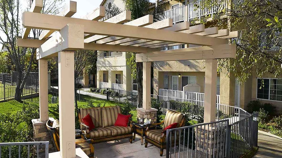 A cozy patio with a pergola, featuring cushioned chairs and surrounded by a residential complex with lush greenery.