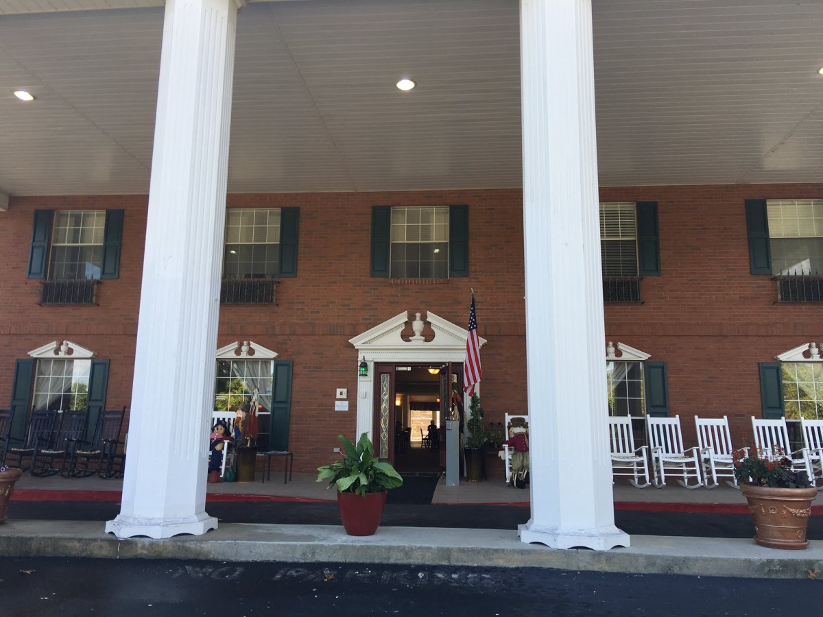 A brick building with large white columns and a porch featuring rocking chairs.